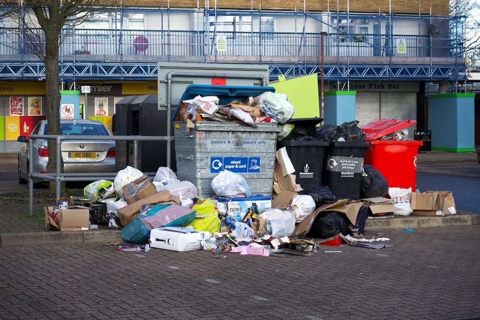 A photograph depicts a collection of variously sized and shaped pieces of discarded furniture and household items placed on a cobbled driveway in front of a brick building. Foremost, there are several