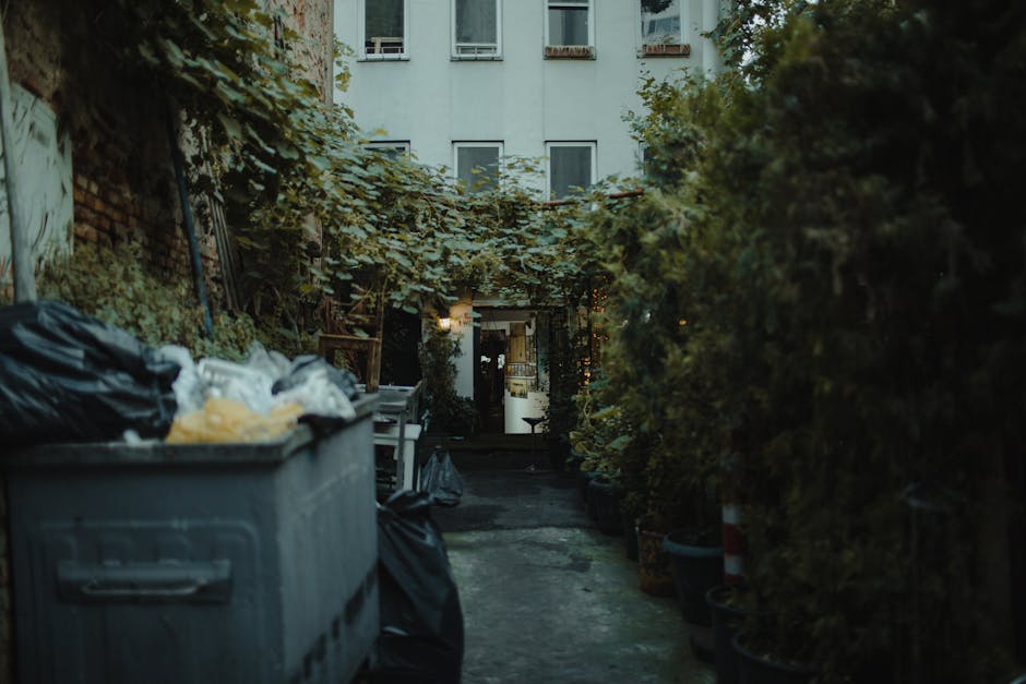 A narrow outdoor alleyway adjacent to a residential building, featuring a large grey plastic rubbish bin on the left side overflowing with black and white garbage bags and other waste materials, some